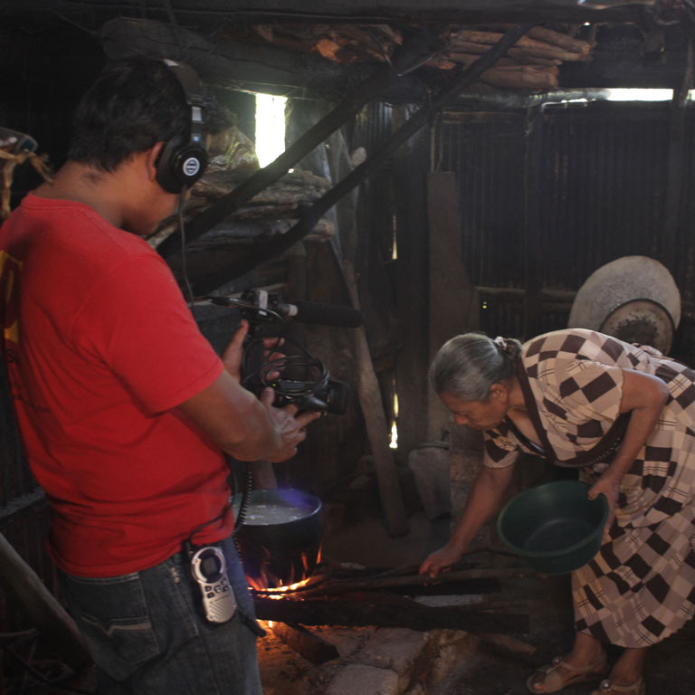 A cameraman records a woman working with a pot over a fire