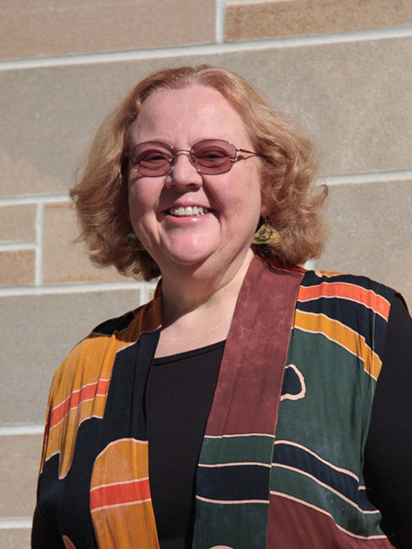 A headshot of Professor Virginia Vitzhum, who wears a multi-colored shirt and poses against a gray limestone wall outdoors.