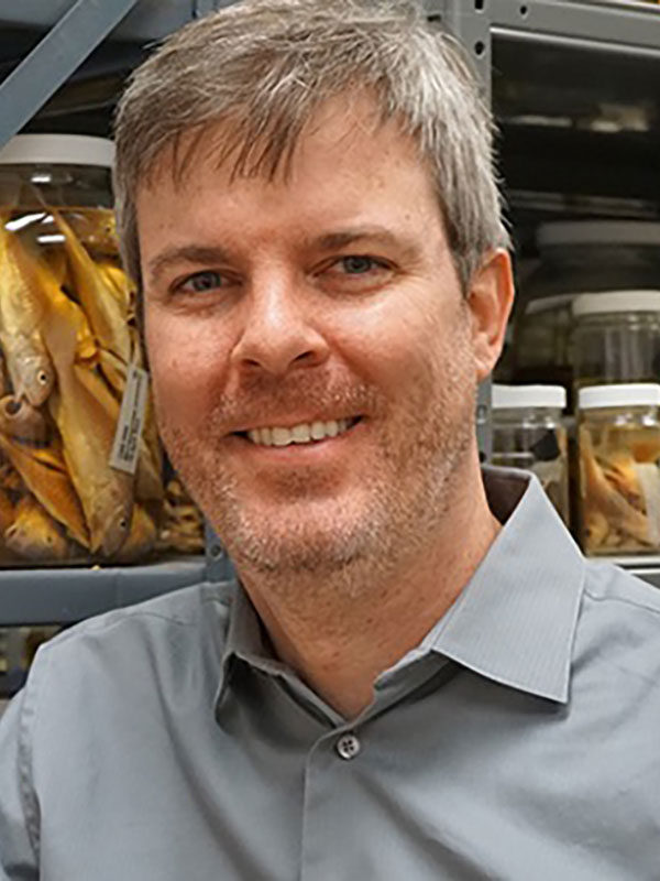 A headshot of Ryan Kennedy, who wears a gray dress shirt and poses in what appears to be a grocery store.